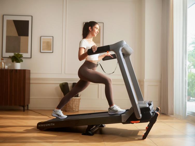 Woman performing a standing lunge stretch beside a KingSmith WalkingPad G5i treadmill in a bright living room.